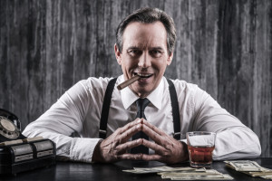 Power is in his hands. Smiling senior man in shirt and suspenders sitting at the table and smoking cigar while lots of money laying near him